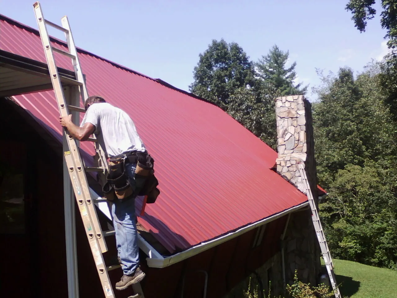 Roofer on a ladder beside a red metal roof and stone chimney — Soffit & Fascia Repair by Roofing Brothers in Palomar Mountain